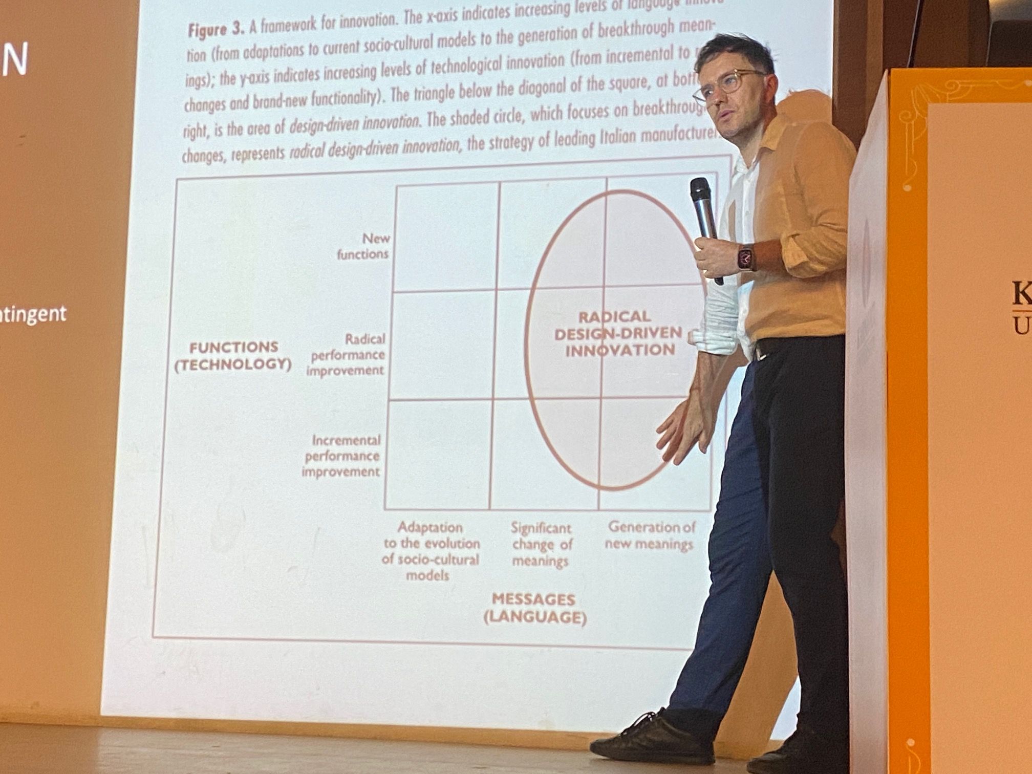 PhD candidate Thomas Luke student stands in front of a projected screen, presenting its information with a microphone in hand